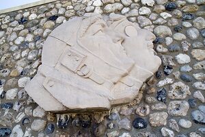 On a stone wall, hangs a stone carving showing two male faces in profile. Together, they are looking upwards and to the right, towards the sky. Both men are wearing aviator helmets and one has an eyepatch over his right eye