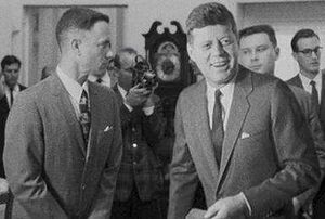 Black-and-white film screenshot showing the main character on the left looking toward another man, President Kennedy, (voiced by actor Jed Gillin), on the right. Kennedy is smiling and looking to his left. In the background, several men are looking in different directions and one is aiming a camera.