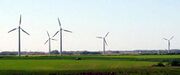 A car drives past 4 wind turbines in a field, with more on the horizon