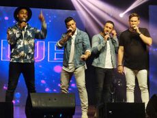 An image of 4 male performers onstage singing to a hand held microphone