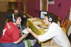 People gathered around a table wearing headphones. The journalist holds the microphone for a physically disabled person to speak