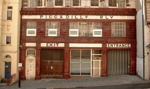 Side elevation of station with the typical red-tiled facade. A pale tiled band with the words "Piccadilly Railway" runs across the top of the second storey and "Exit" and "Entrance" are displayed above shuttered doorways.