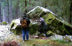 Two people with their backs to the viewer stand in front of a large boulder in the middle of woodland. One of the figures is pouring a liquid onto the ground.