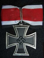 A silver framed black cross that has arms which are narrow at the center, and broader at the perimeter. In the middle of the cross is a swastika, an equilateral cross with its arms bent at right angles. On the lower arm of the cross are the number 1939 engraved. The cross is connected to a ribbon with a silver clip. The ribbon has a red central stripe, flanked in white and with a black edge stripe.