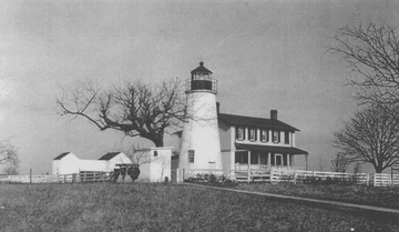 A white lighthouse and attached two-story house on a hill. There is farmland around the house. A large black walnut tree is also pictured behind the station.