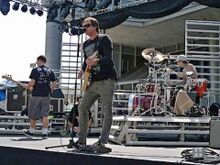 A black-haired man plays a guitar while another man plays a drum kit in the background.