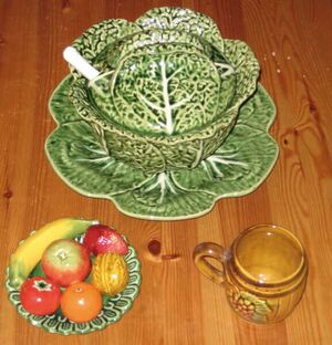 top center: a cabbage-shaped tureen; bottom left: decorative plate with ceramic fruits and walnut; bottom right: barrel-shaped mug with grape motif